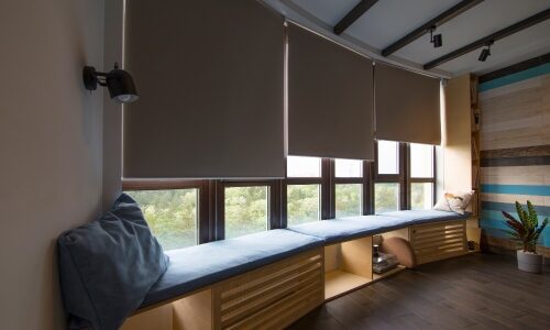 Motorized shades in the interior. Automatic roller blinds beige color on big glass windows. Remote Control Shades are above the windosill with pillows. Summer. Green trees outside. Olibra study
