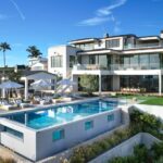 A modern white villa with a large pool, surrounded by palm trees and lounge chairs, against a clear blue sky.