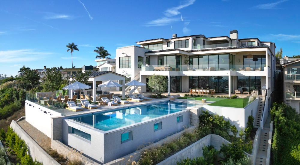 A modern white villa with a large pool, surrounded by palm trees and lounge chairs, against a clear blue sky.