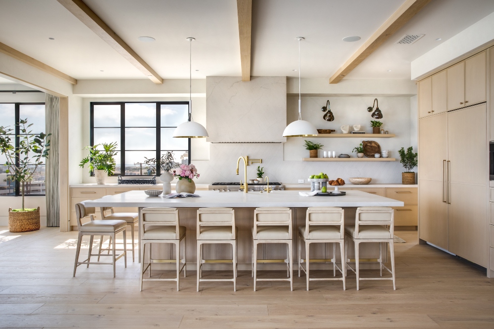 A modern kitchen with a large island, cream-colored cabinets, and stylish bar stools. Lush plants add greenery