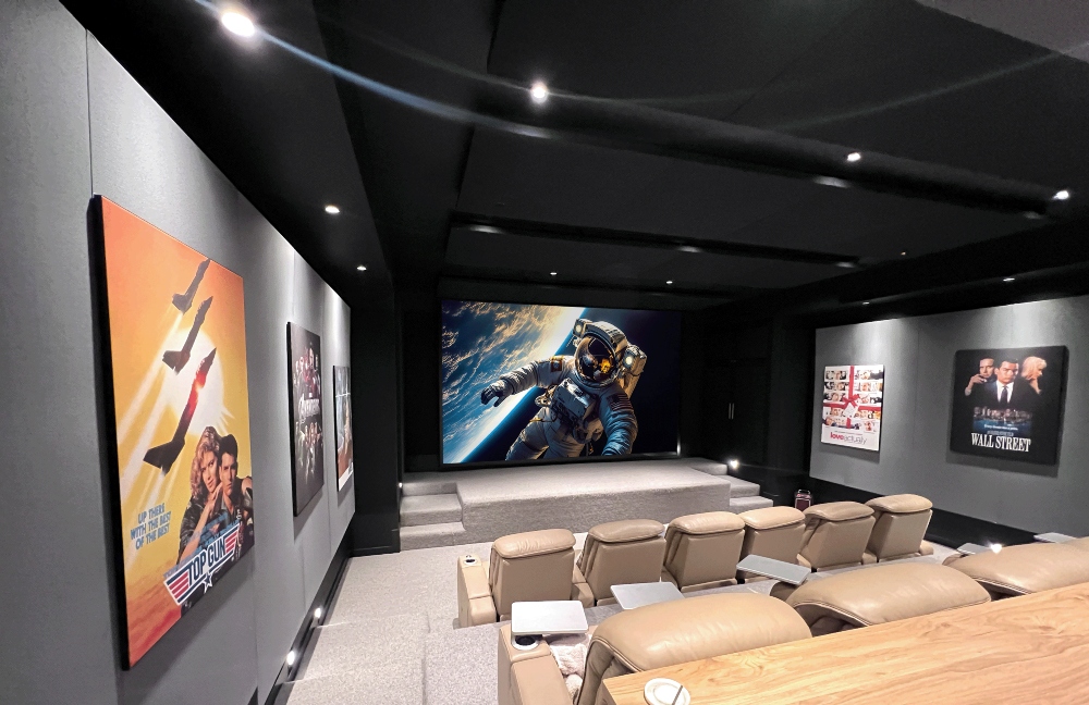 Luxury home theater with tiered leather recliners, dark acoustic walls, a large screen displaying an astronaut image, and framed movie posters including ‘Top Gun’ and ‘Wall Street’ on the side walls.