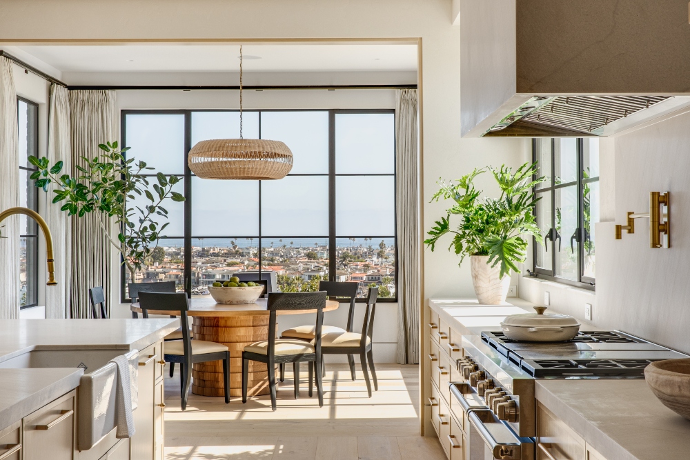 Bright contemporary coastal kitchen and dining area with large black-framed windows, light wood cabinetry, indoor plants, a round wood dining table, and ocean views in the background.