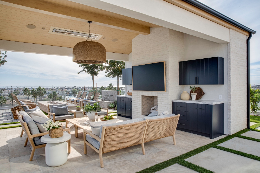A contemporary coastal outdoor living space with a seating area, TV, and view of palm trees and water.