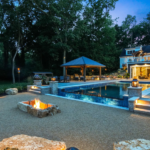 Infinity-edge pool illuminated at night with landscape lighting reflecting across the water in a scenic riverfront backyard.