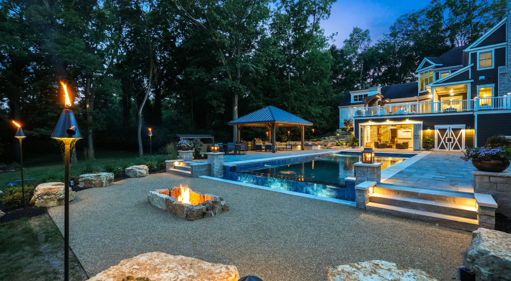 Infinity-edge pool illuminated at night with landscape lighting reflecting across the water in a scenic riverfront backyard.
