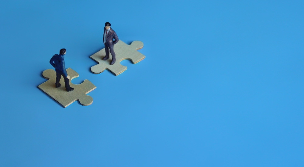 Two men standing on puzzle pieces meant to represent the business transaction of mergers and acquisitions concept for consolidation within an industry, particularly within CI, to represent the analysis provided in CE Pro's weekly playback news segment for the custom integration industry.
